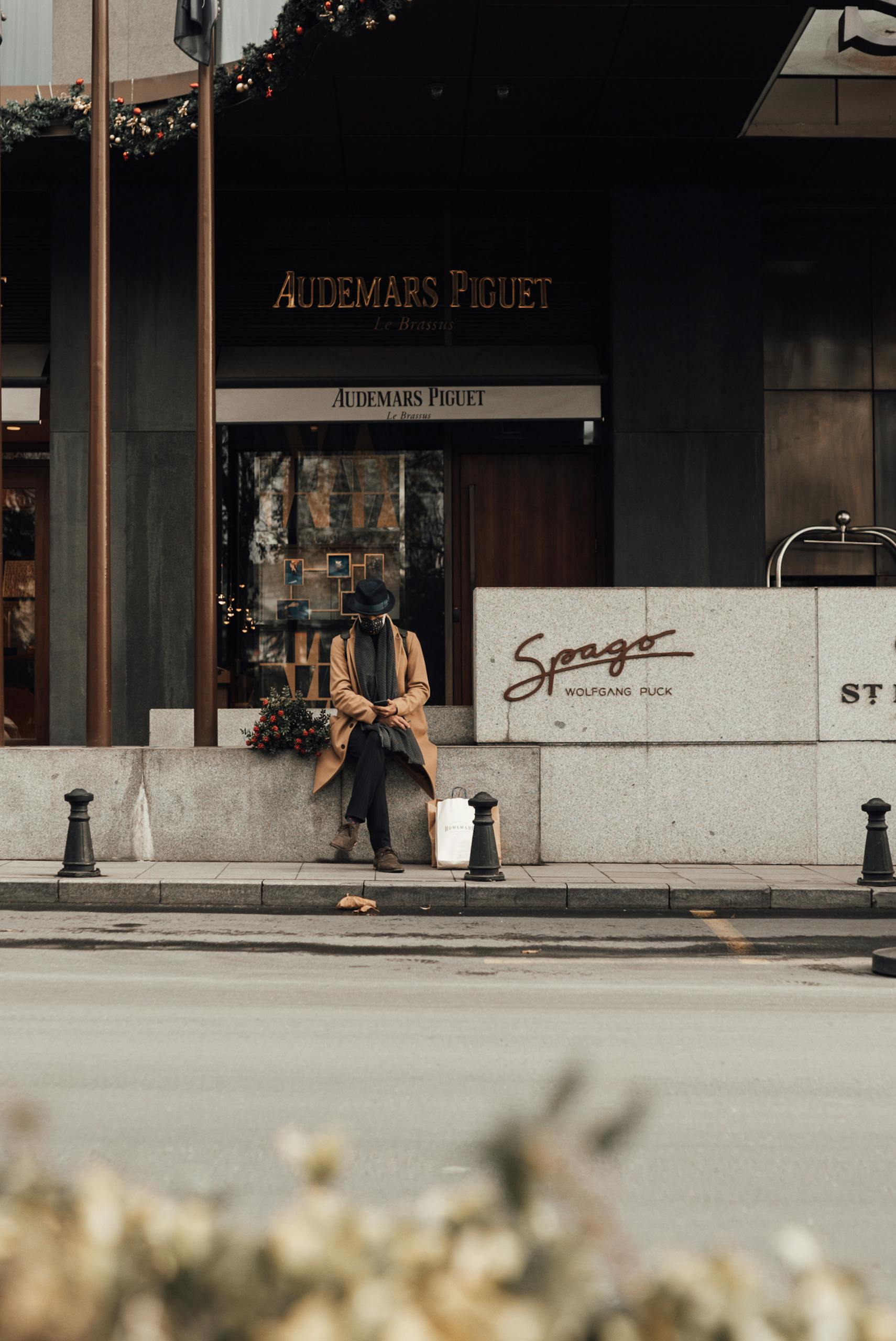 Masked person sitting outside a luxury store with winter attire on a city street.